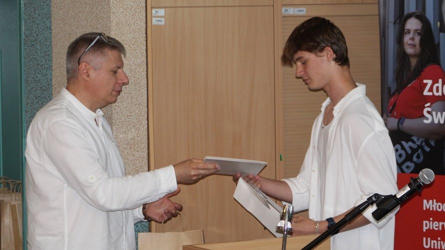 Dwaj mężczyźni podczas oficjalnego wręczenia dokumentu. Mężczyzna po lewej stronie, ubrany w białą koszulę, wręcza dokument młodszemu mężczyźnie po prawej stronie, który również jest ubrany w białą koszulę. W tle znajdują się drewniane szafki oraz fragment czerwonego roll-upu z napisem "Zdolny Uczeń - Świetny Student". 