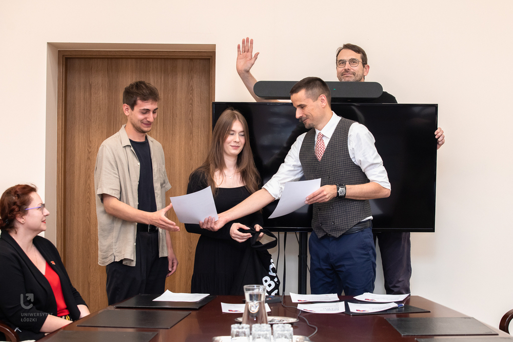 Moment wręczania dyplomów od prof. Krzysztofa Pabisa. Na drugim planie Rektor, prof. Rafał Matera machający do fotografa zza dużego ekranu telewizora. Moment wręczania dyplomów od prof. Krzysztofa Pabisa. Na drugim planie Rektor, prof. Rafał Matera machający do fotografa zza dużego ekranu telewizora.