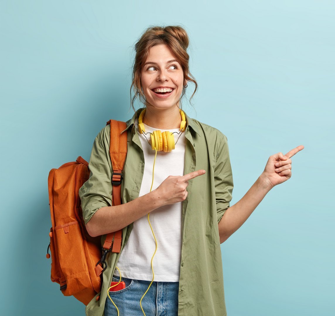 Positive European female teenager uses modern technologies for entertainment, points aside on free space, dressed in loose shirt and jeans, carries backpack, promotos something, poses indoor Student