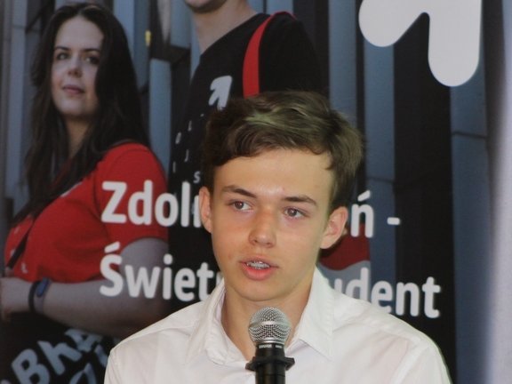 Młody mężczyzna przemawiający do mikrofonu. Jest ubrany w białą koszulę i wygląda na skoncentrowanego. W tle widoczny jest plakat z napisem "Zdolny Uczeń - Świetny Student" 