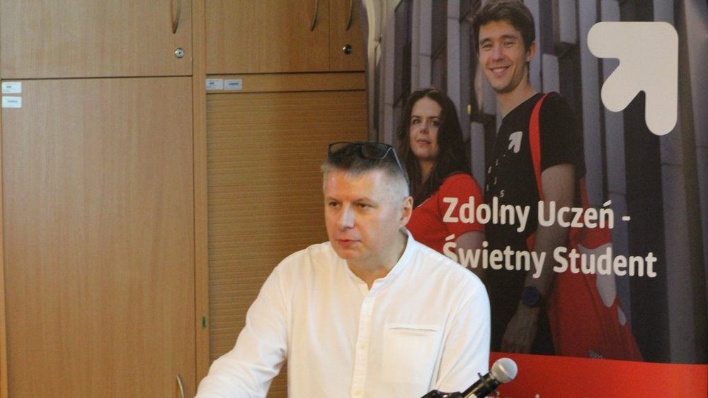Mężczyzna stojący przed biurkiem, ubrany w białą koszulę, wygląda na skupionego. Za nim widać duży plakat z napisem "Zdolny Uczeń - Świetny Student. Na biurku przed mężczyzną znajduje się mikrofon, co sugeruje, że może być w trakcie przemowy. 