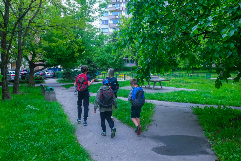 Children going to school through a park. They have backpacks.