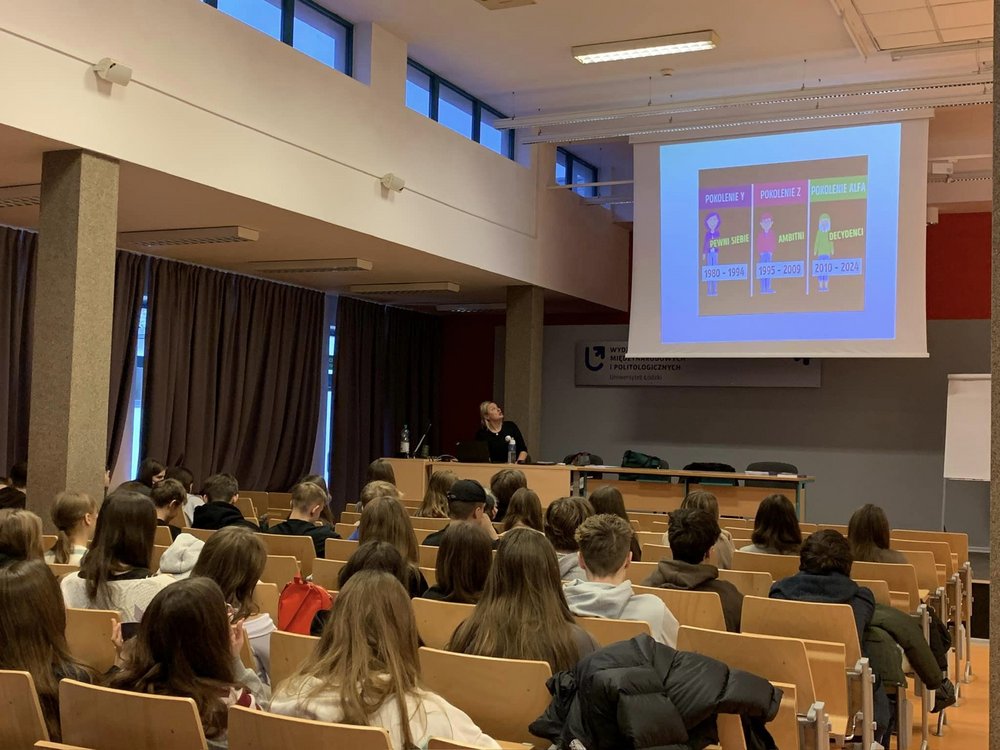 The photo shows a group of people in a lecture hall. There is a projector in the room with a multimedia presentation. A lecturer is standing at the projector.