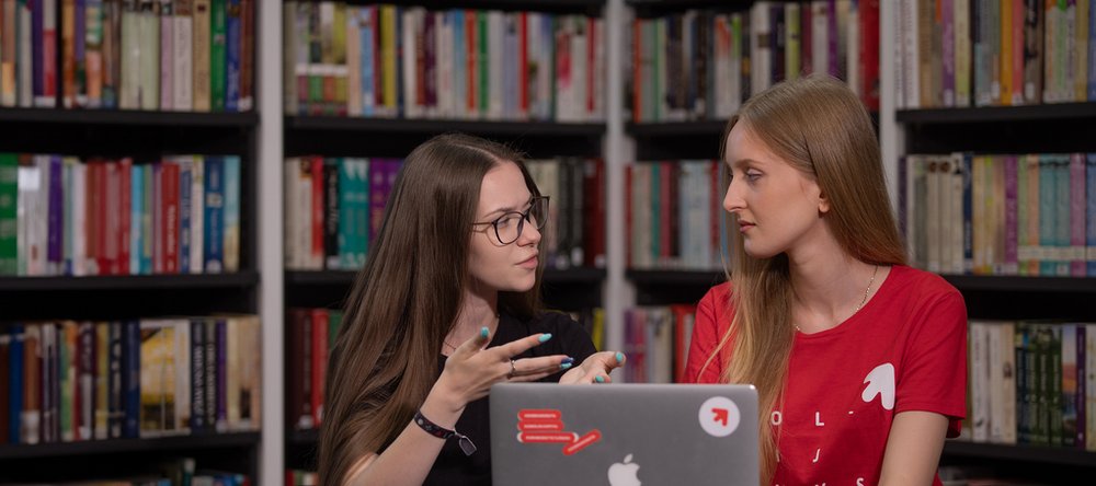 a colourful photo showing two female students consulting together on the implementation of a project against the background of bookshelves