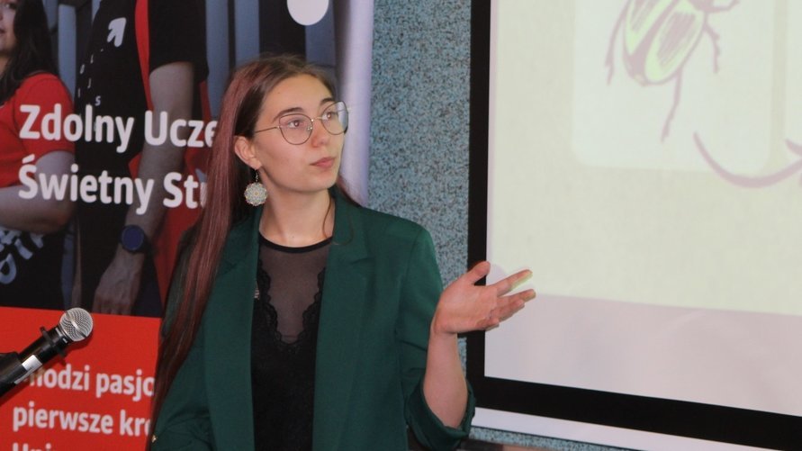 Młoda kobieta, ubrana w zieloną marynarkę i czarną bluzkę, a na twarzy ma okulary prowadzi prezentację. W tle widoczny jest ekran projekcyjny, na którym wyświetlana jest ilustracja, oraz czerwony roll-up z napisem "Zdolny Uczeń - Świetny Student".