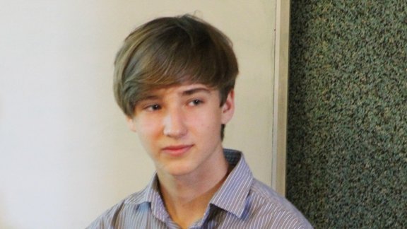 Młody mężczyzna o jasnych włosach, ubrany w koszulę w delikatne paski. Patrzy w bok z delikatnym uśmiechem na twarzy. Tło stanowi ściana o teksturze przypominającej tapetę w ciemnym kolorze. 