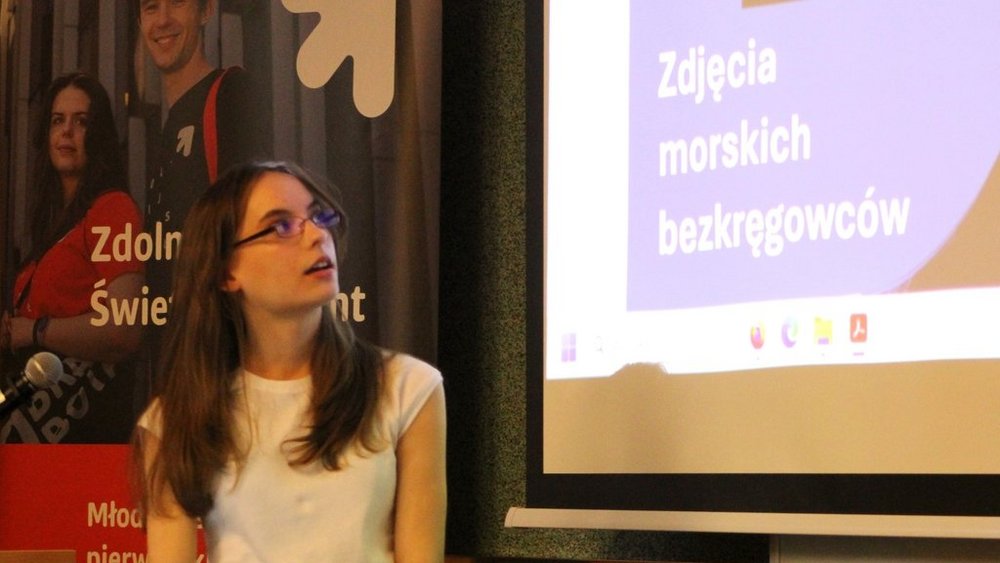 Na zdjęciu znajduje się młoda kobieta prowadząca prezentację. Jest ubrana w białą bluzkę i okulary. Patrzy w górę, na ekran projekcyjny, na którym widnieje slajd z napisem "Zdjęcia morskich bezkręgowców". W tle po lewej stronie widoczny jest plakat z napisem "Zdolny Uczeń - Świetny Student" 