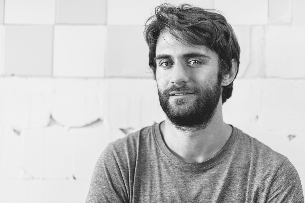 czarnobiałe zdjęcie przedstawiające uśmiechniętego młodego mężczyznę z brodą patrzącego wprost w obiektyw