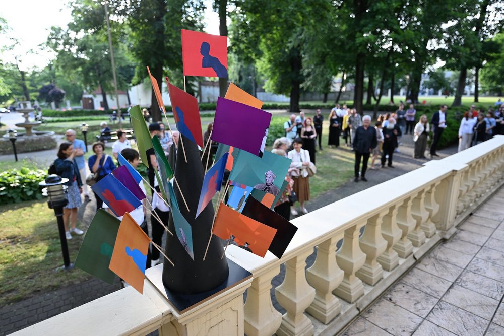 The photograph shows a park filled with people. It is spring, and the trees are green. In the foreground, a concrete balustrade is visible, with a stand displaying colourful bunting placed on top. In the background, the aforementioned people can be seen walking.