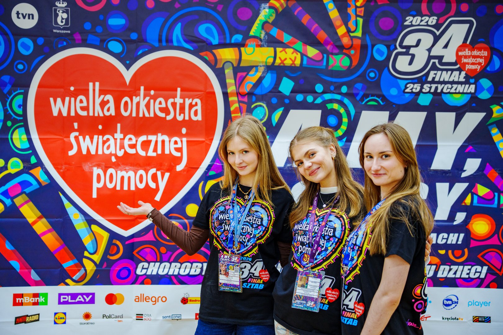 3 młode kobiety ubrane w czarne koszulki z napisem Wielka Orkiestra Świątecznej Pomocy