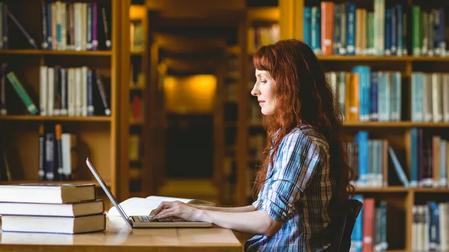 A young woman sitting in the foreground working on a laptop; a library collection of books is in the background